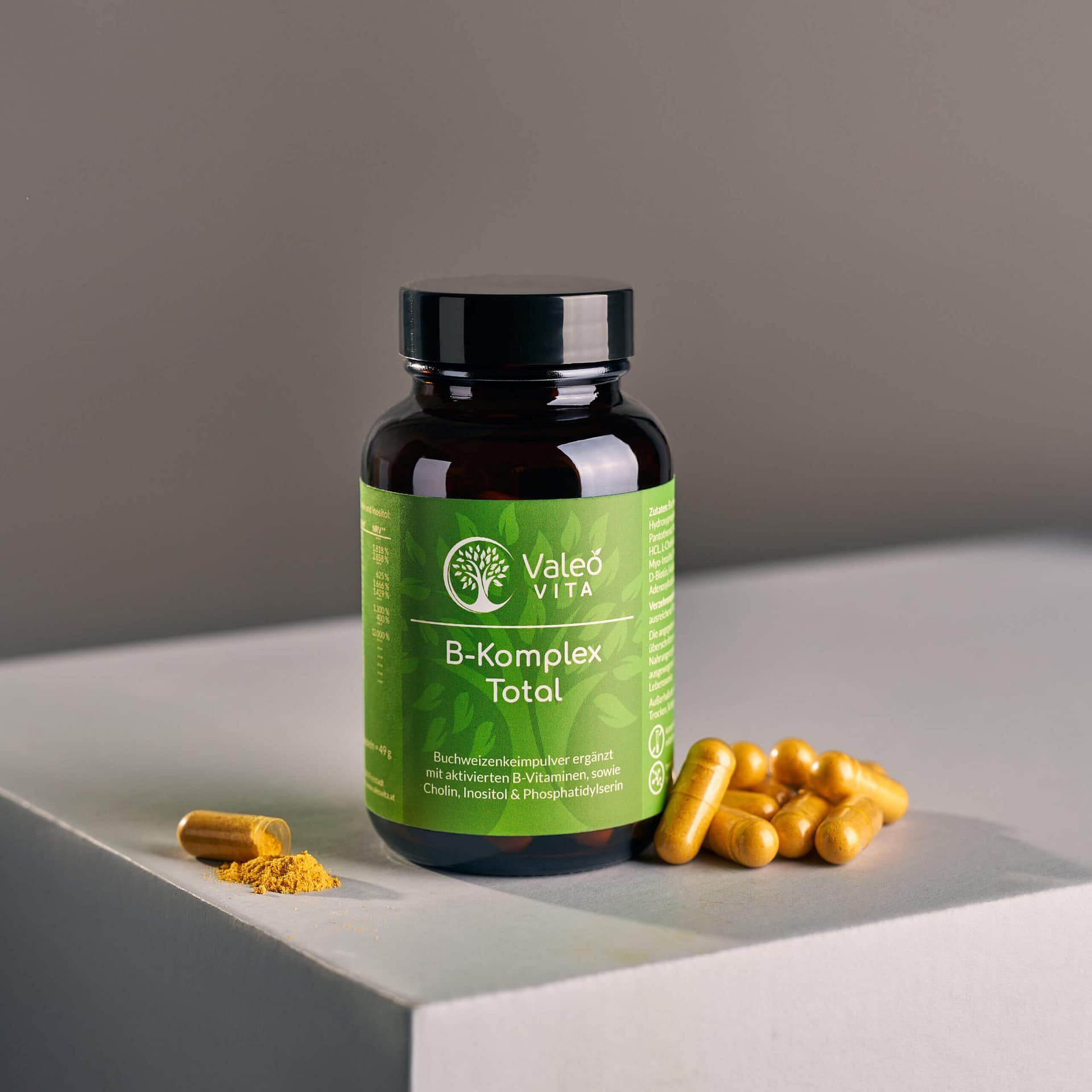 Dose mit Valeo Vita B-Komplex Total Kapseln, gelbe Kapseln und etwas Pulver auf hellem Podest