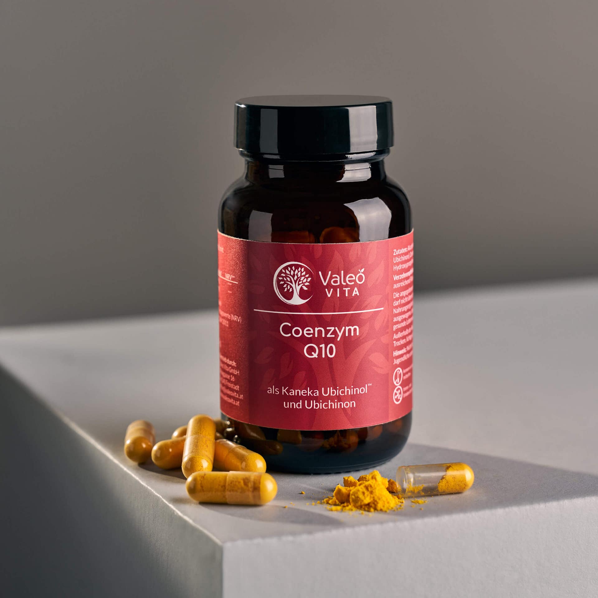 Valeo Vita Coenzym Q10 Kapseln in Braunglasflasche mit Kapseln und Q10-Pulver auf grauem Podest