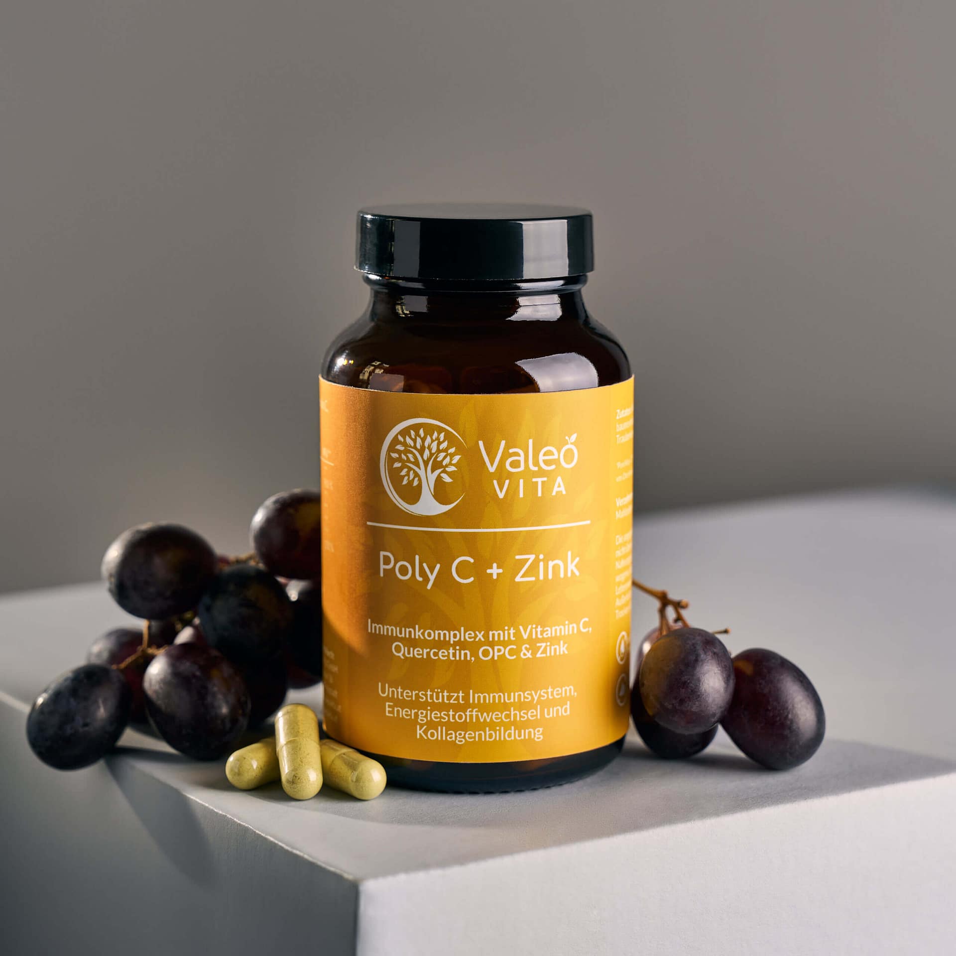 Dose mit Valeo Vita Poly C plus Zink Kapseln neben Trauben und Kapseln auf hellem Podest